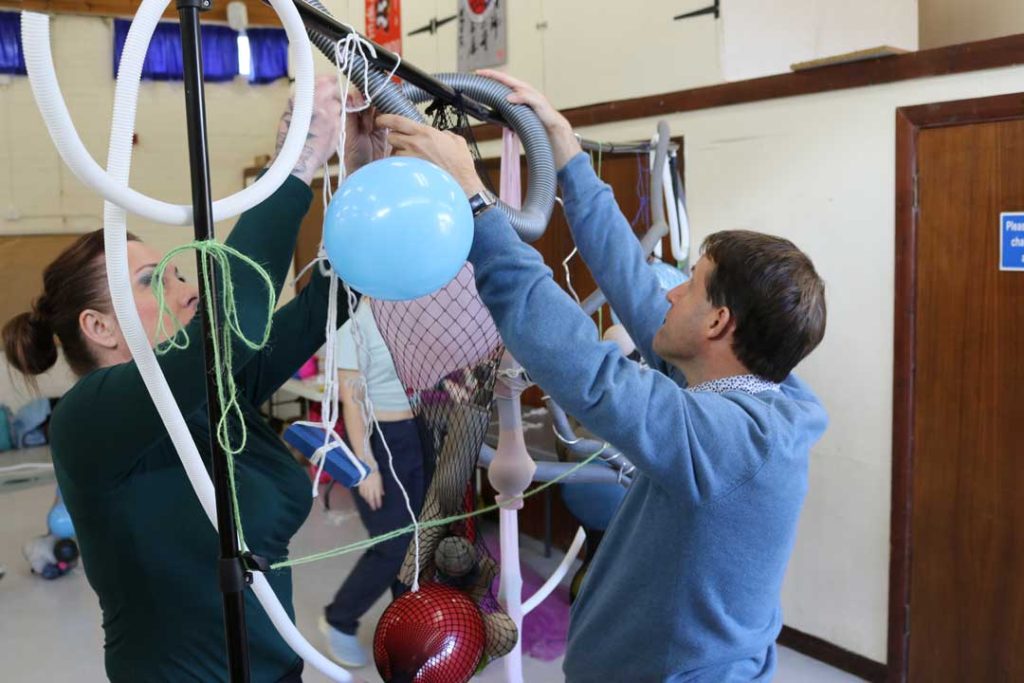 A man and woman are smiling while affixing vibrant balloons and assorted items to a structure