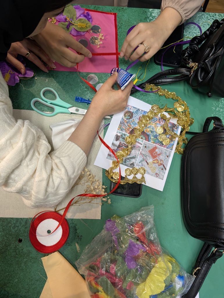Overhead view of several people’s hands working together at a craft table, arranging ribbons, coins, artificial flowers, and trims on pink fabric, with scissors, thread, and reference images scattered around.