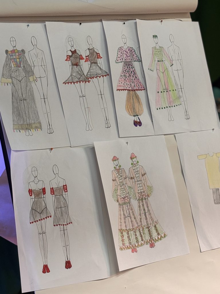A wall of hand-drawn fashion sketches showing multiple garment designs on mannequin figures, coloured with pencils and featuring embroidered details, patterned fabrics, and traditional-inspired silhouettes.